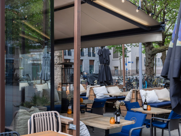 Overdekt horecaterras met terraszonwering, houten tafels en comfortabele blauwe zitplaatsen.