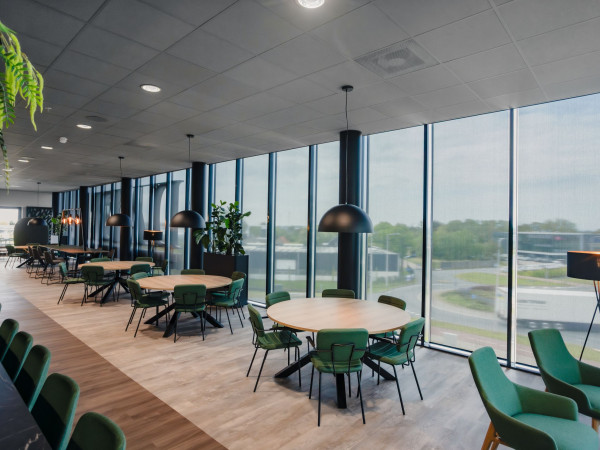 Bedrijfsrestaurant met groene stoelen en binnenzonwering langs de glazen buitengevel