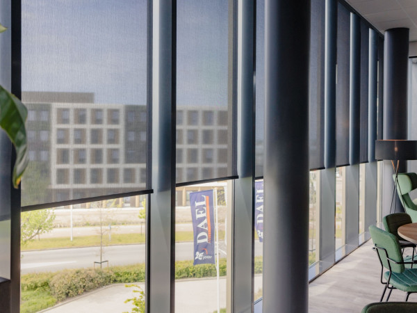 Gedeeltelijk geopende rolgordijnen langs de glazen gevel van een moderne bedrijfskantine.